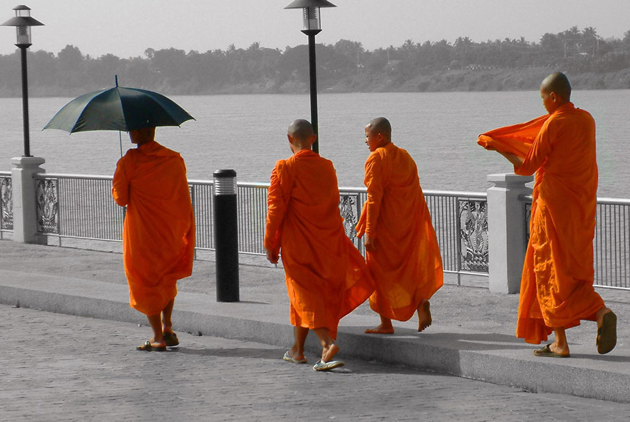 4-monks-in-orange-robes-walking-on-bridge-along-river-one-under-black-umbrella